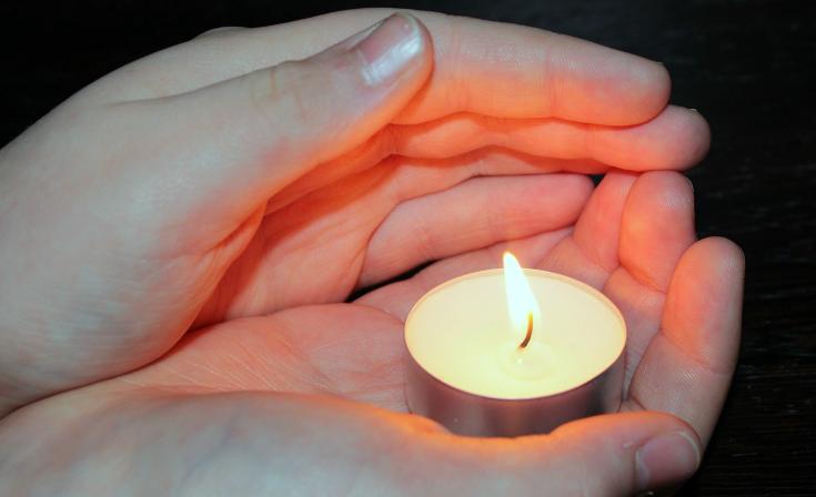 A tealight candle held in the palm of someone's hand as their other hand shelters the flame.